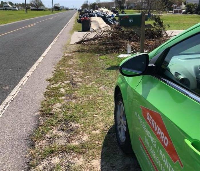 Image shows green SERVPRO car along a residential street with debris piled on the side of the road.